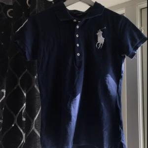  - Marinblå Ralph Lauren piké med vita detaljer som som den stora trean på höger axel. Det är strl M men skulle mer säga att den är som XS/XXS köparen betalar för frakt :)