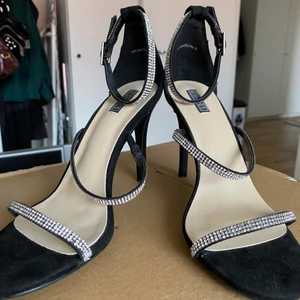 Nelly klackar med glitterremmar - Svarta klackar från Nelly med tre remmar med stenar. Använda en gång! Jättefina men tyvärr lite stora för mig! Klackarna är 10 cm!👠