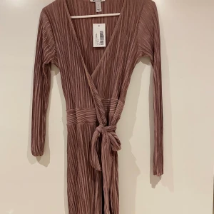 Rosa plisserad klänning, oanvänd, S - Rosa plisserad klänning, omlott med knyt, Nelly. Oanvänd med lappar kvar. Storlek small. 