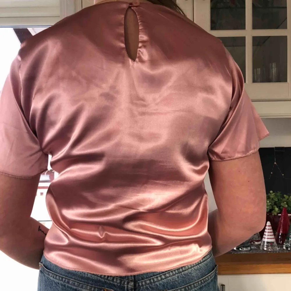 Snygg t-shirt från Nelly i ett fint rosa blankt tyg. Med snygg knytning fram samt knapp bak i ryggen. Knappt använd så i fint skick. Köparen står för frakt🥰. T-shirts.