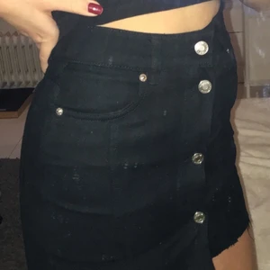 Svart jeanskjol med slitning - Svart jeanskjol från Weekday med knappar framtill och fickor fram och bak. Avklippt nertill. Använd fåtal gånger. Skriv för fler bilder. Buda med minst 10 kr höjning!💖
