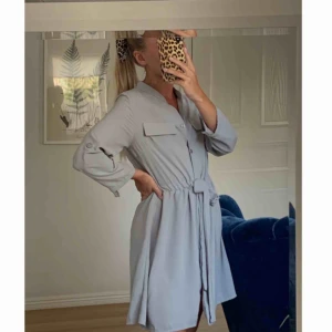  - Ljusblå/grå långärmad klänning med knytbart band i midjan & knappar längs hela framsidan. Knappar på armarna också ifall man vill kavla upp ärmen. Köpt i Australien, i storlek UK8 vilket motsvarar en 36a. Frakt tillkommer.