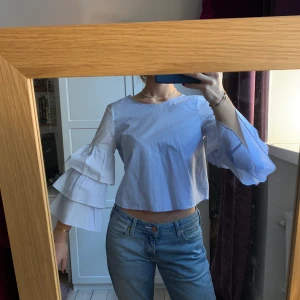 Ljusblå Zara blus med volangärmar - En blus med vackra volangärmar och öppen rygg med knytning. Blusen går i en ljusblå skala där ärmarna även har små ränder (den första bilden visar bäst färgen på blusen). Använd en gång och säljer därför. Storlek S. Hör av er vid frågor💙