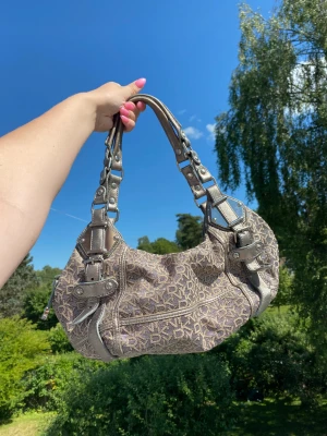 Glittrig DKNY väska - En så fin brungrå glittrig väska från DKNY! 🤩🙌Vid ett ställe har tråden gått upp lite (se sista bilden) samt att handtagen ser använda ut men annars är den i fint skick!!🥰 Luktar dessutom lite rök men håller på att vädra den!☺️