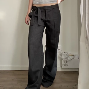 Grå lågmidjade kostymbyxor med skärp - Grå lågmidjade kostymbyxor med pressveck, avtagbart skärp, fejk-fickor bak och riktiga fickor på sidan. Innerbenslängd: 85cm överkant omkrets: 93cm