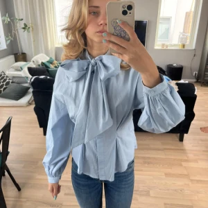 Söt ljusblå knytblus - Ljusblå knytblus med stor rosett fram. Insydda veck bak och fram för fin smal siluett. Ballongärm.