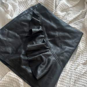 En svart skinn kjol med volang på framsidan. Du knäpper med en dragkedja på baksidan. Använd fåtal gånger men ser ut som ny.