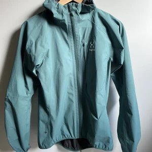 Haglöfs funktionsjacka  - Obefintligt använd. Figursydd kvinnomodell. Coretex material som är vattenresistent. Fin grön/grå färg. Ärm innerlängd 57 cm. Storlek S.