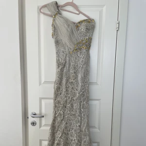 Klänning - Exklusiv silver/grå/beige spetsklänning med pärlor och stenar. One shoulder och släp. I använt skick och behöver tvättas därav priset. 