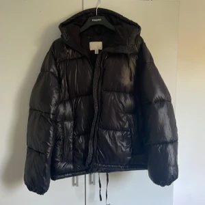 Svart glansig vinterjacka  - Här är en varm vinterjacka som är i väldigt bra skick, använd halva vinern förra året, går även att dra åt längst ner och även vid luvan, storlek xs men väldigt stor i modellen. Dunjacka 