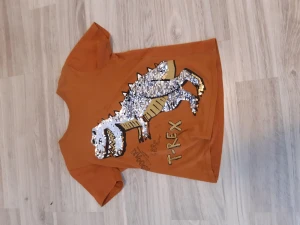 T-shirt orange med t-rex - Nyskick T-shirt. Dinosaurien har glitter som man kan vända på så att man får 2 stilar