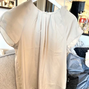 Blus - Säljer en bege t-shirt blus i ett glansigt tyg, med dragkedja i bak. Tröjan är använd fåtal gånger, så i ett mycket fint skick.  Färg: Bege Storlek: S