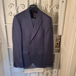 Jamonte kostym blå – Tiger of Sweden – OANVÄND - Material: 98 % ull, 2 % elastan. Kostym i ullstretch. Smal passform. Något bredare slag. Knäpps med två knappar Helfodrad. Tordon byxor, låg midja och smala ben. Premiummaterial från det berömda italienska väveriet Marlane. Resegarderob medföljer.