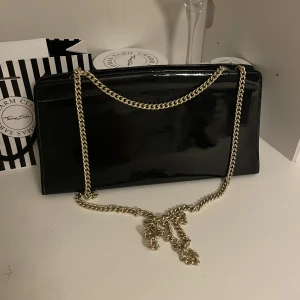 Svart glansig axelremsväska med guldig kedja - Kuvertväska, liten väska med guldig kedja. Perfekt storlek till fester eller middagar. Får plats med mobil, nycklar, kort och läppglans m.m. Den är i väldigt bra skick och har knappt använts. Jag köpte den för cirka 600kr. Skriv för fler frågor eller bild