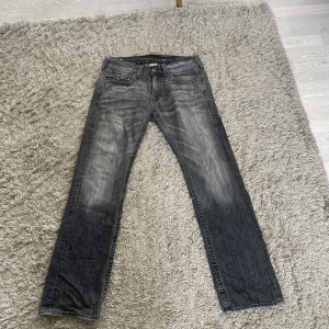 Gråa True Religion Ricky Jeans - Ett gammalt par gråa true religion jeans med rak passform. Har inte använts mycket men saknar en av knapparna på vänster bakficka.  Storlek 33. 