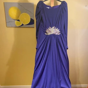 Mörklila balklänning  - Mörklila balklänning med långa ärmar och vackert strassmycke framtill. Snyggt draperad i midjan bäde fram och bak.   Använd en gång, som ny.  Design: Trina Lewis & Marjon Couture, London. Tillverkad i England  Storlek: 14 i Storbritannien, 12  i USA, 40