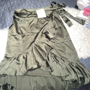 Grön satin omklottkjol - Olivgrön omlottkjol i satin med volanger längs kanten.  Stl. XS köpt från Göteborg Shoko Design Använder den inte längre så säljer den för 300kr+fraktpris (nypris 500kr)