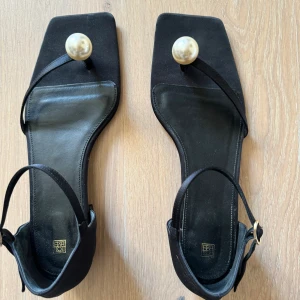 Sandaler ”the pearl flat black” från Toteme  - Sandaler i svart satin med vacker pärla (ej äkta pärla). Skorna har bara använts en enstaka gång och är nästan som helt nya. Storlek 41, men jag skulle säga att de är mer som 40.  Skokartong med påse kommer med. Kvitto från Toteme finns.