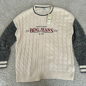 Beige stickad tröja från Urban Outfitters - En beige stickad tröja från Urban Outfitters med kabelstickat mönster och texten 'BDG JEANS' i rött på framsidan. Tröjan har långa ärmar i grått och ribbstickade muddar vid ärmslut och nederkant.