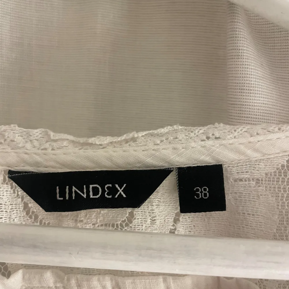 En vit topp från Lindex med spetsdetaljer på ärmarna och längs med knäppningen framtill. toppen har korta ärmar och en knytning i midjan för en justerbar passform. Den är dekorerad med knappar framtill.. Puserot.