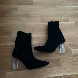 Svarta stövletter med genomskinlig klack - Ett par eleganta svarta stövletter med spetsig tå och genomskinlig klack. Skorna har en tight passform och är tillverkade i ett syntetiskt material. Aldrig använda pga fööör stora, storlek US8 eller oxå 40/41