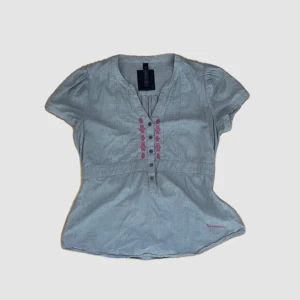 Grå blus med broderi - En blå blus med korta ärmar och broderade detaljer i rosa på framsidan. Blusen har knappar framtill och ett knytband i midjan för en justerbar passform.