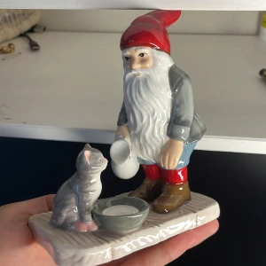 Keramikfigur av tomte med katt - En detaljerad keramikfigur föreställande en tomte med röd luva, grå jacka, blå byxor och bruna stövlar. Tomten håller en vit kanna och står bredvid en grå katt som sitter vid en skål. Figuren är målad i naturliga färger och har en glansig yta.