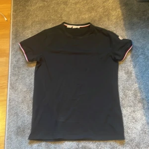 Svart t-shirt från Moncler - En svart t-shirt från Moncler med korta ärmar. T-shirten har en enkel design med en liten logotyp på vänster ärm och röd-vit-randiga detaljer vid ärmsluten. skriv om due intresserad 