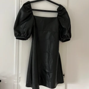 Svart klänning med puffärmar - En elegant svart klänning med puffärmar och en fyrkantig urringning. Klänningen har en kort längd och är tillverkad i ett glansigt material. Baksidan har en dekorativ snörning.