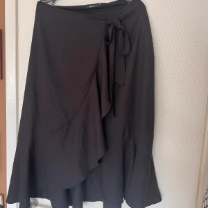Svart omlottkjol - Fin och bekväm omlottkjol med volang. Tyget är tunt men inte genomskinligt. Stl 38 men passar som en storlek 40. Knäpps med knapp men kan göras tightare med snörningen 💓 Går till knäna på mig som är 174 🌷