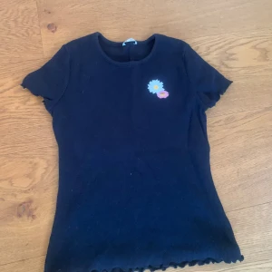 Svart ribbad topp med blommig detalj - En svart ribbad topp med korta ärmar och en liten blommig detalj på bröstet. Toppen har en vågig kant vid ärmslut och nederkant.