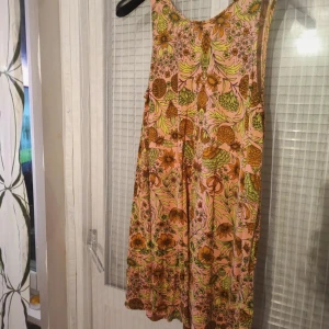 Blommig klänning - En blommig klänning med ett färgglatt mönster i nyanser av rosa, grönt och gult. Klänningen är ärmlös och har en lös passform, vilket gör den bekväm och luftig.