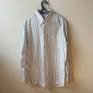 Lacoste Skjorta - En vit-randig skjorta från Lacoste med logga på bröstfickan | Storlek 40/Medium | Mycket fint skick 👍  Kolla in min profil för fler skjortor, pikétröjor och annat till bra pris! Fler varor kommer inom kort 💸