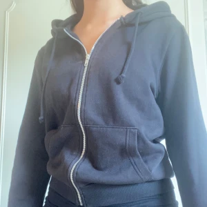 Svart hoodie med dragkedja - En bekväm svart hoodie med dragkedja framtill. Hoodien har en justerbar huva och en stor ficka på magen. Den är långärmad och har ribbade muddar vid ärmslut och nederkant.