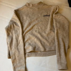Beige tröja från FB Sister - En beige tröja från FB Sister med texten 'united' på bröstet. Tröjan har långa ärmar och en hög krage.