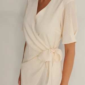  Vit klänning med knytdetalj - Kort omlottklänning från NA-KD i gräddvit. Helt oanvänd i storlek 40. Originalpris: 699:- Lite större i bysten än vad jag hade gillat men materialet är lätt och bekvämt mot kroppen, har en insydd underklänning. 