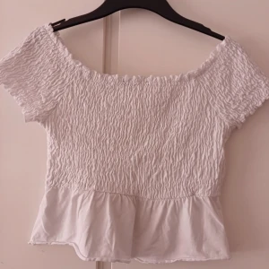 Vit off-shoulder topp - En vit off-shoulder topp med korta ärmar och en rynkad design på överdelen. Toppen har en volangdetalj vid nederkanten. Använd men ser ut som ny!