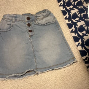 Ljusblå jeanskjol från Charanga - En ljusblå jeanskjol från Charanga, designad i Spanien. Kjolen har tre knappar framtill, två fickor fram och två fickor bak. Den har en rå kant nedtill som ger en avslappnad look.