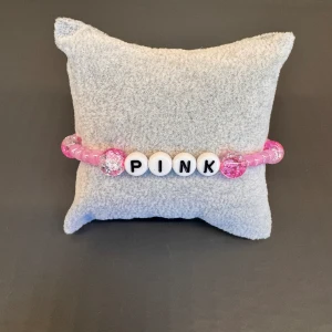 Pärlarmband  - Egentillverkat pärlarmband med rosa pärlor och med texten ”PINK” med svart-vita pärlor. Är gjort med gummitråd och är ca 17 cm långt.