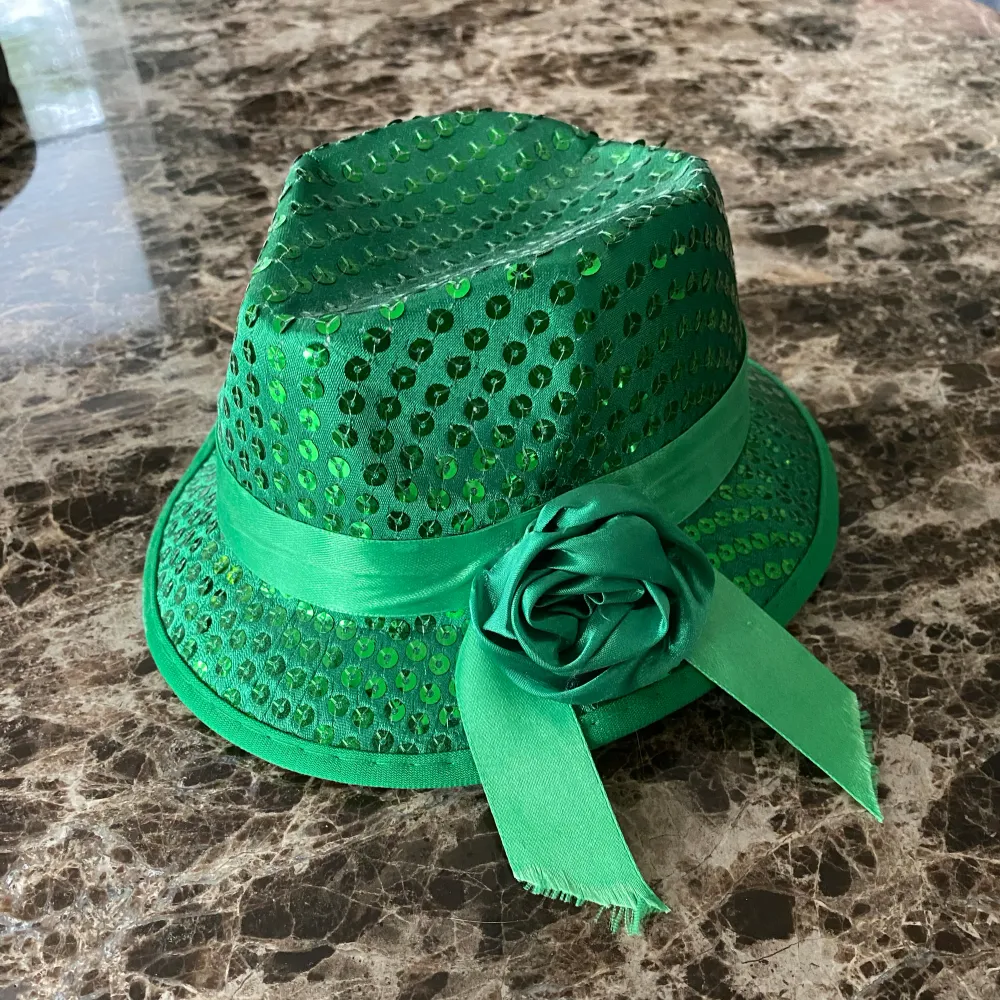 En grön hatt med gröna paljetter på med ett grönt band format som en ros. Är i perfekt skick (nästan aldrig använd). Saknar ingen paljett. Perfekt för fester. Enkel att sätta på, med silver klippor. Ca 20cm i längd, ca 17,5cm i bred + 10 cm i höjd.. Accessoarer.