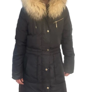 Hollies jacka  - Svart, figursydd dunjacka i längre modell från Hollies. Luva med avtagbar pälskrage. Dold mudd i ärmslut I storlek 34. 