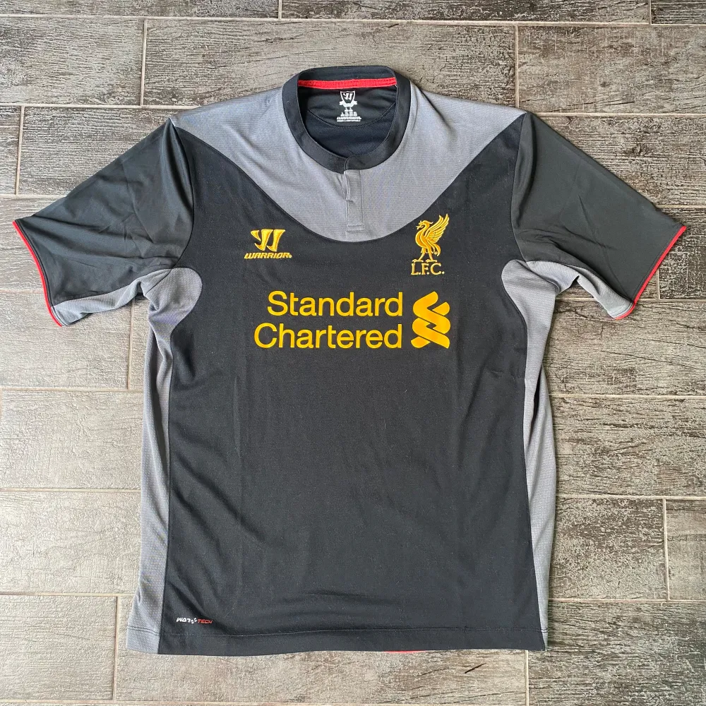 En svart och grå fotbollströja från Warrior med Liverpool FC:s emblem och sponsorlogotypen 'Standard Chartered' i gult på framsidan. Tröjan har korta ärmar med röda detaljer och en rund halsringning.. T-shirts.