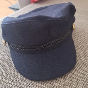 Mörkblå keps med flätad detalj - En mörkblå keps med en flätad detalj och guldknappar på sidorna. Kepsen har en skärm och är tillverkad i ett mjukt material.