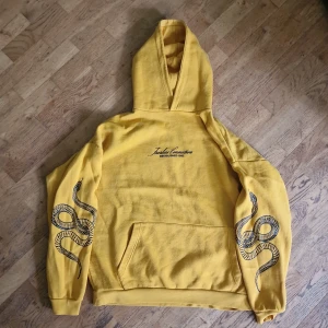 Gul hoodie från Black Squad - En gul hoodie från Black Squad med en svart ormtryck på ärmarna. Hoodien har en känguruficka framtill. Den är i en oversized passform. Den är hel men rätt använd.