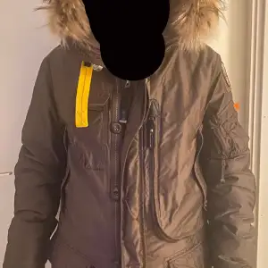 Brun, kraftig dunjacka från Parajumpers med flera praktiska framfickor och en innerficka med dragkedja.Jackan har även ett avtagbart dunfoder.Jackan har en snygg avtagbar krage i naturpäls runt luvan samt det ikoniska gula kabinhaken.Dragkedja Stl stor M