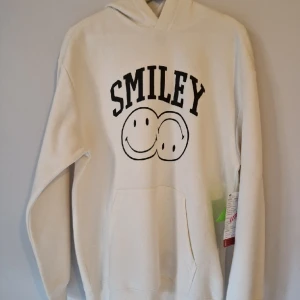 Vit hoodie "Smiley" strl M, oanvänd - Vit hoodie från H&M med svart tryck "Smiley". Storleken är Medium, relaxed fit (dvs oversized, så det är en stor medium). Hoodien är oanvänd och är förmodligen från H&M's provkollektion nov 2021, osäkert om den kom ut i produktion.