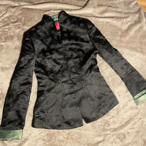 Jacka med kinesiska tecken - En tunn, kavaj liknande jacka i svart med gröna ärm ändar och grönt inne i jackan. Silkes liknande material. Storlek S. För liten för mig (176) i både armar och axlar.  Bra kvalitet.