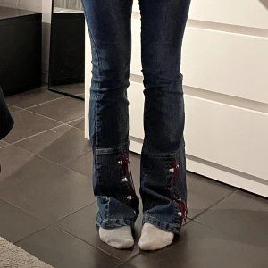 Lågmidjade jeans - Unika jeans med coola, vinröda detaljer. Jättecoola, knappt använda och  lågmidjade. Passar någon med 34/36 i storlek. Rakt över midjan: 38 cm. Innerbenslängd: 81 cm. 