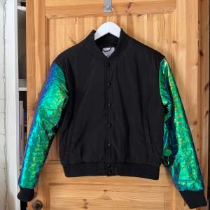 Svart bomber med skimrande ärmar i prassligt material. Lite oil slick. Märkt M men passar XS-S bäst då den är liten i storleken. Personen på bilden är 176 cm