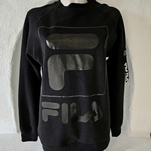 Svart Fila sweatshirt crewneck med tryck - Svart sweatshirt crewneck med svart glansigt tryck från Fila, fick på armen med snygg detalj. Som ny. Vita luddet på bilden är från en annan tröja. Den är nu kemtvättad.